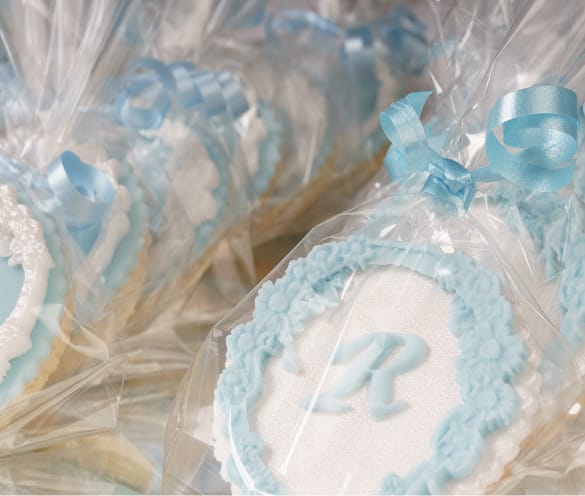 Biscotti artigianali decorati con glassa bianca e azzurra, caratterizzati dalla lettera "R" al centro e confezionati singolarmente in sacchetti trasparenti chiusi da un nastro azzurro.
